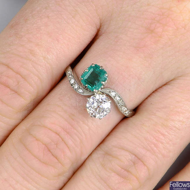 An early 20th century platinum emerald and old-cut diamond two-stone crossover ring, with diamond shoulders.